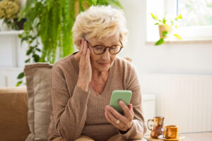 Sad senior woman wearing casual brown clothes sitting on sofa at home and using mobile phone.